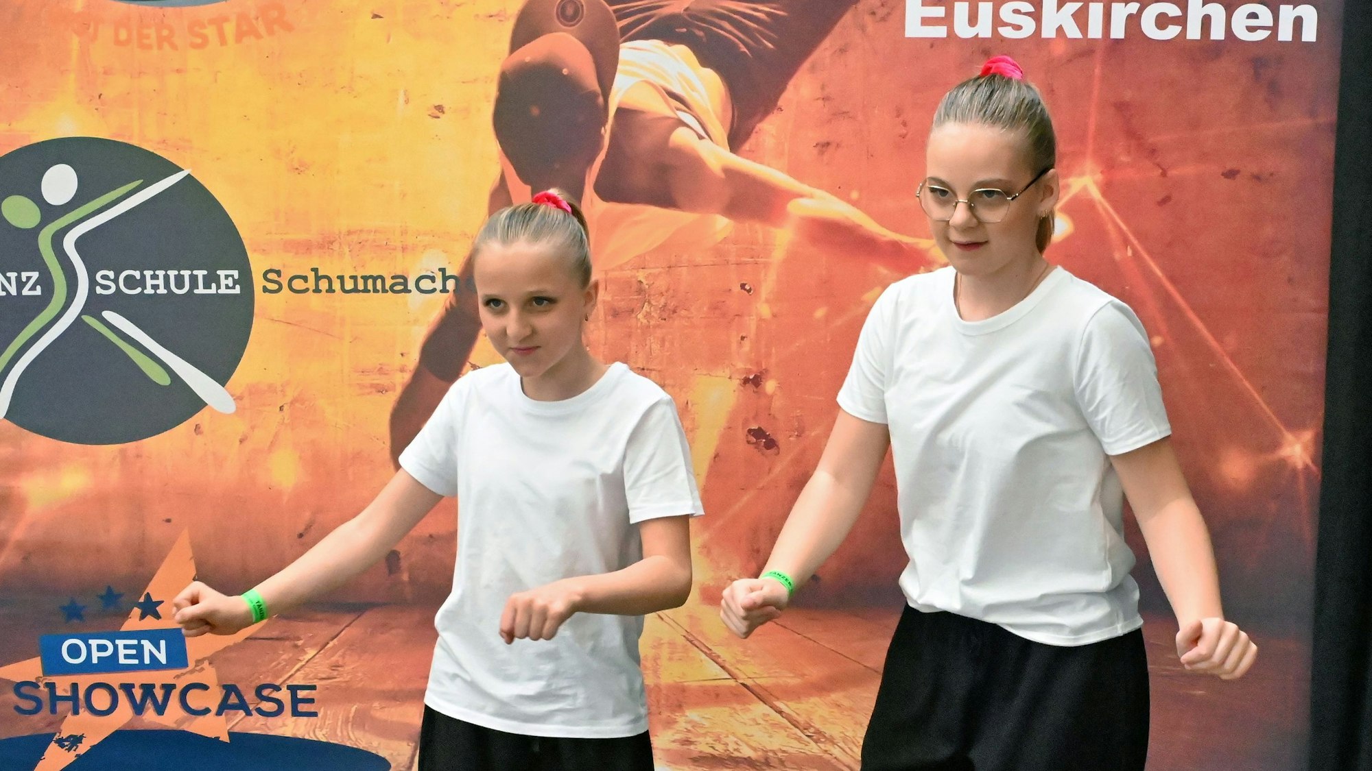 Zwei Mädchen in schwarzen Hosen und weißen Shirts machen Tanzbewegungen.