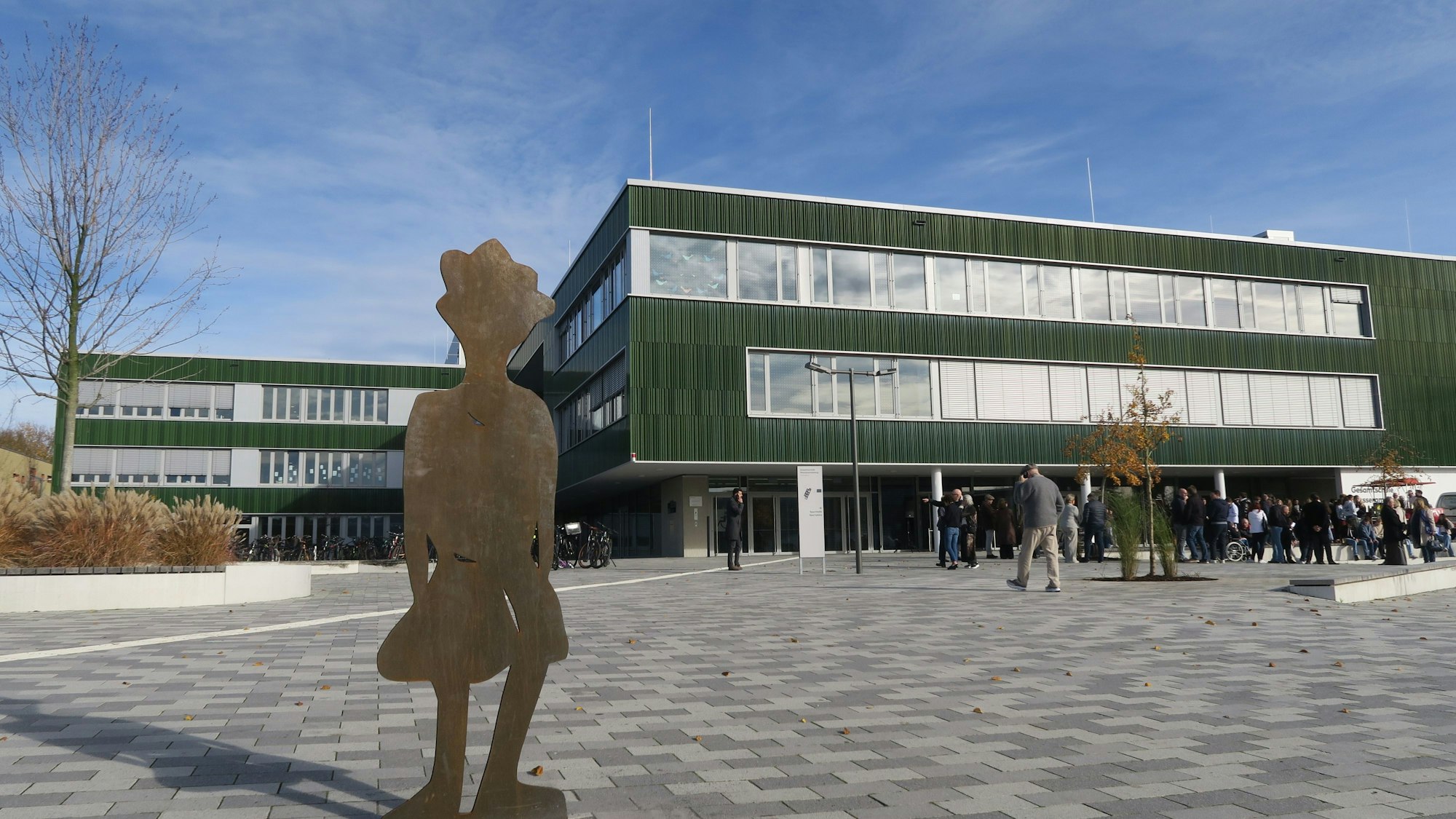 Ein dreistöckiger grüner Flachbau ragt hinter einem großen Platz auf. Davor stehen viele Jugendliche und eine Figur aus Cortenstahl.