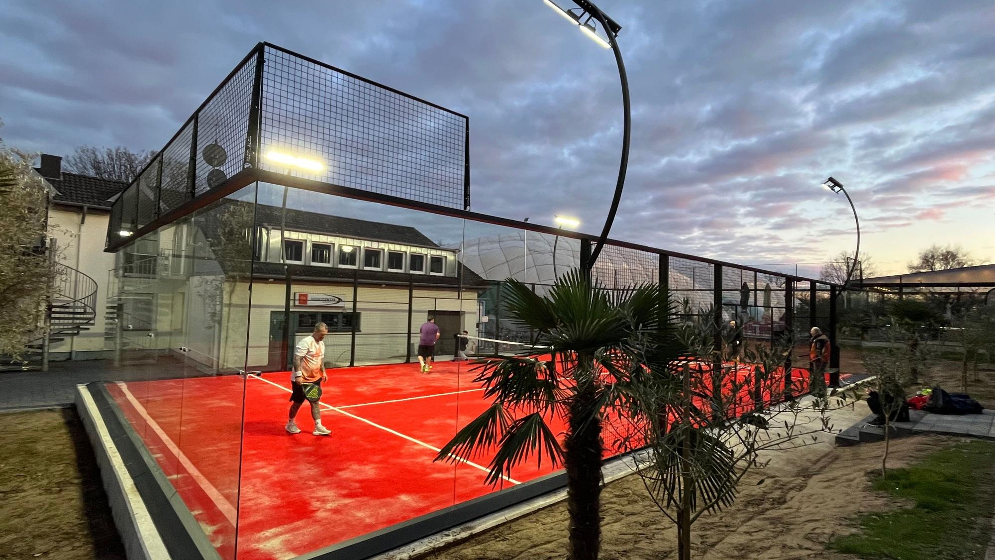 Eine Art Mini-Tennisplatz mit Glaswänden steht vor einem Vereinsheim.