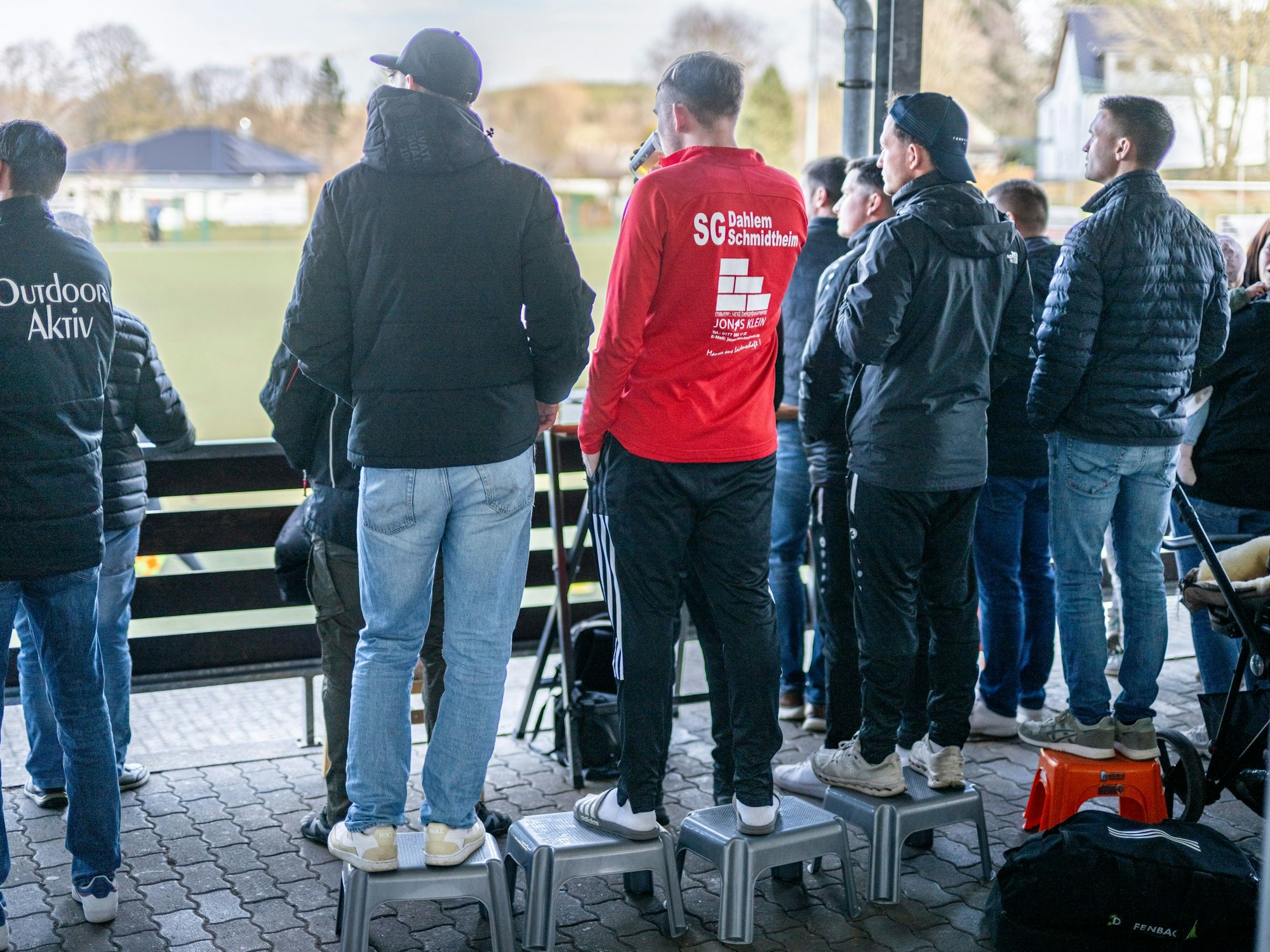Das Bild zeigt einige Fans, die auf Hockern stehen und so das Fußballspiel verfolgen.