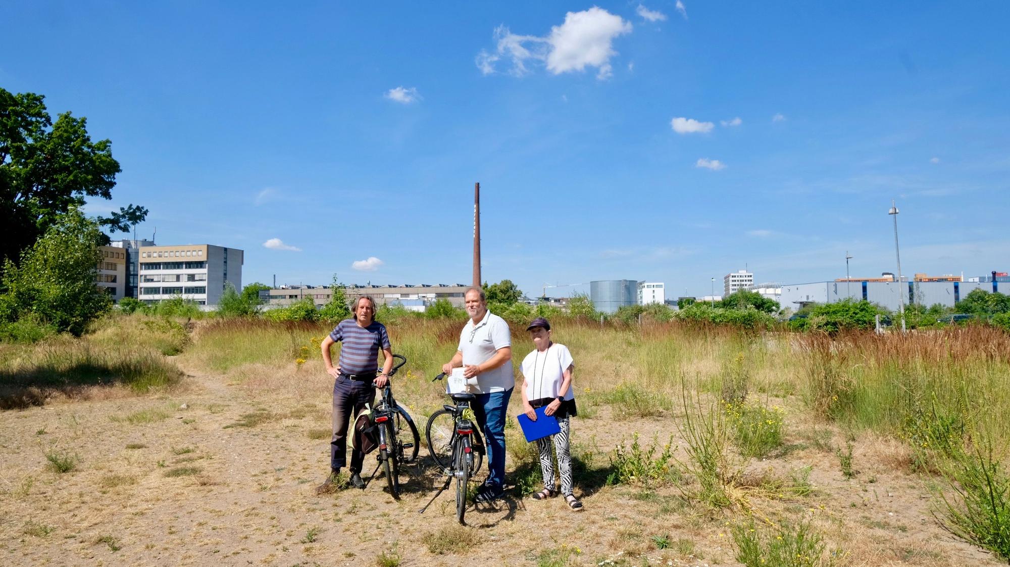 Zwei Männer mit Fahrrad und eine Frau stehen auf einer großen Brachfläche.