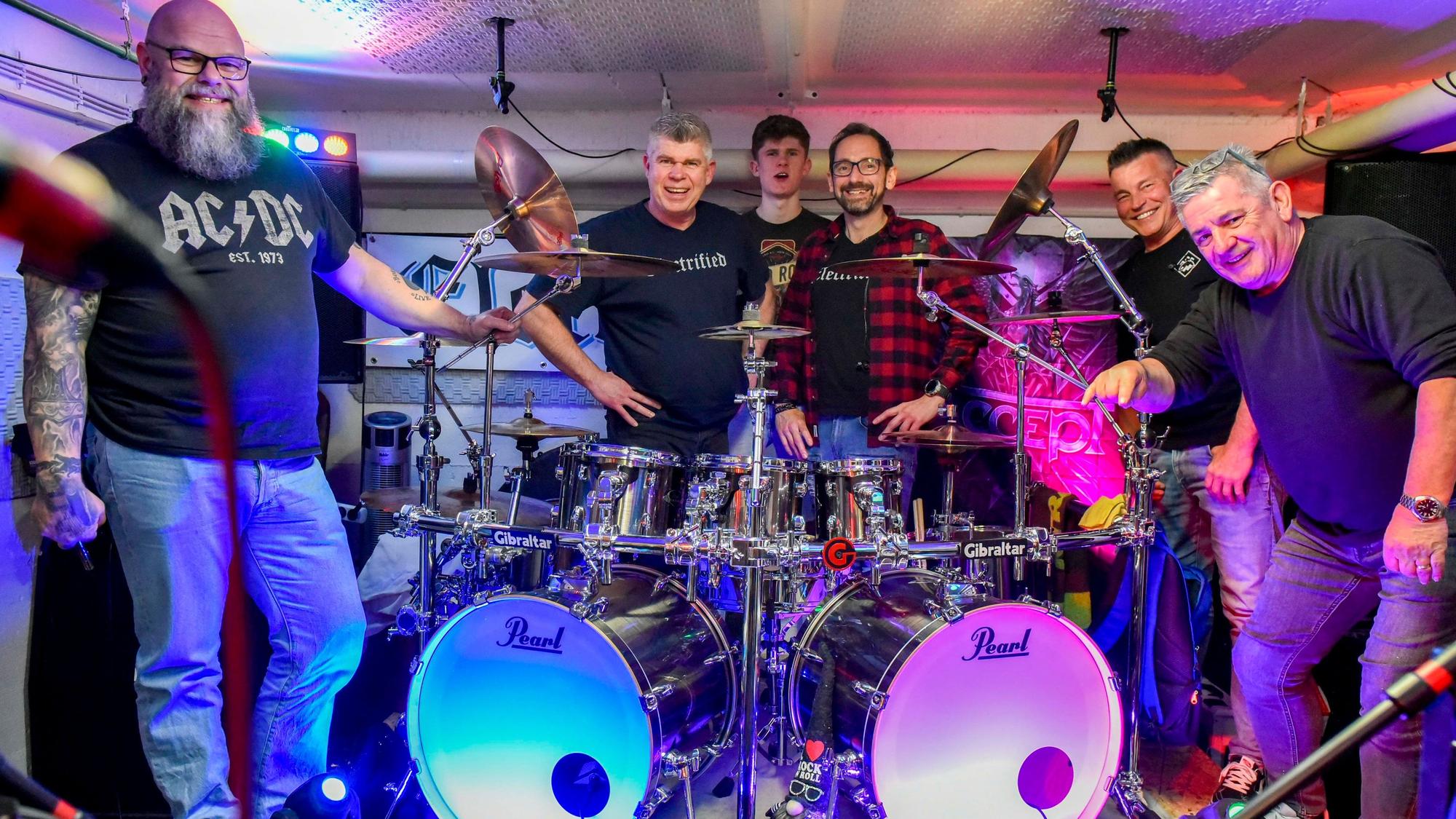 Auf dem Bild sind die Musiker der Frechener Band Electrified zu sehen, die im Proberaum um ein Schlagzeug versammelt sind.