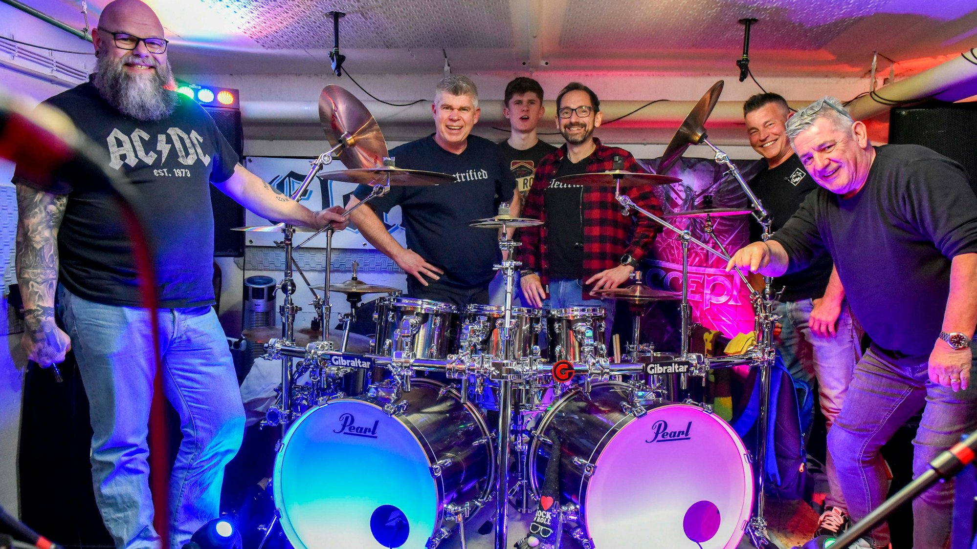 Auf dem Bild sind die Musiker der Frechener Band Electrified zu sehen, die im Proberaum um ein Schlagzeug versammelt sind.