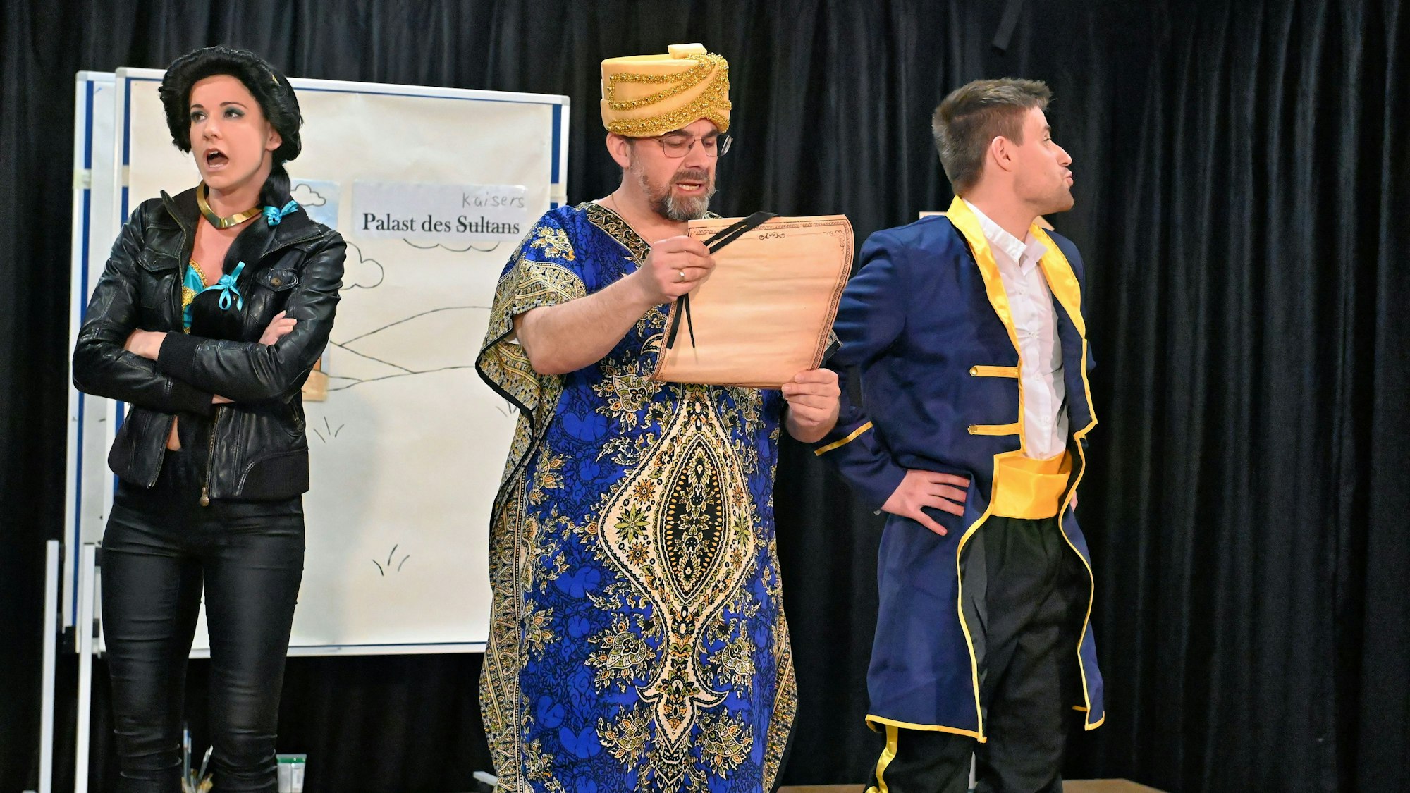 Zwei Männer und eine Frau stehen in Kostümen auf der Bühne.