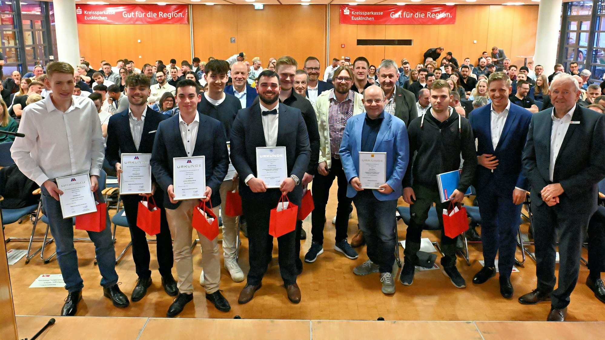 Eine Gruppe Männer, einige haben Urkunden in der Hand. Hinter ihnen ist Publikum zu sehen.