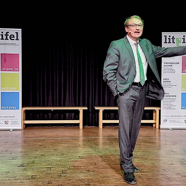 Autor und Papst-Experte Andreas Englisch steht auf einer Bühne und spricht in ein Mikrofon. Im Hintergrund sind zwei Lit.Eifel-Aufsteller zu sehen.