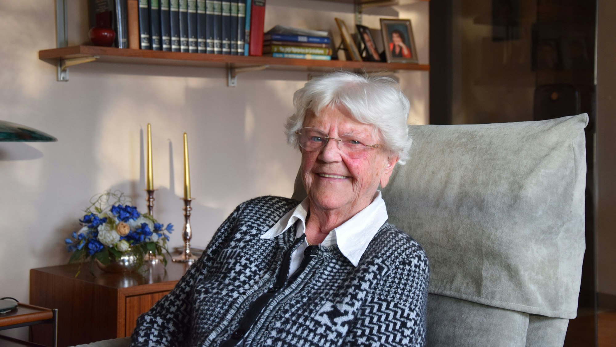Eine Frau mit weißem Haar und Brille sitzt in einem Sessel und blickt lachend in die Kamera.