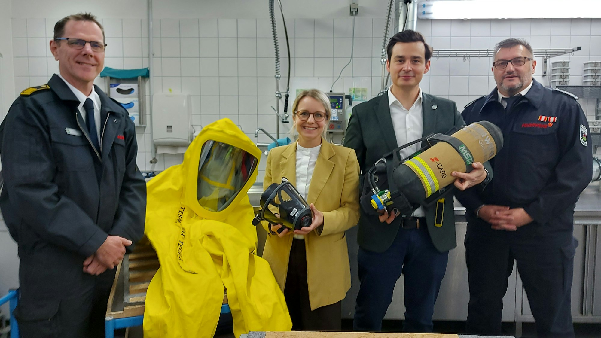 Drei Männer, davon zwei in Feuerwehruniform, und eine Frau im gelben Blazer stehen in einer Werkstatt neben einem gelben Ganzkörperschutzanzug und halten eine Atemschutzmaske und eine Sauerstoffflasche in der Hand.