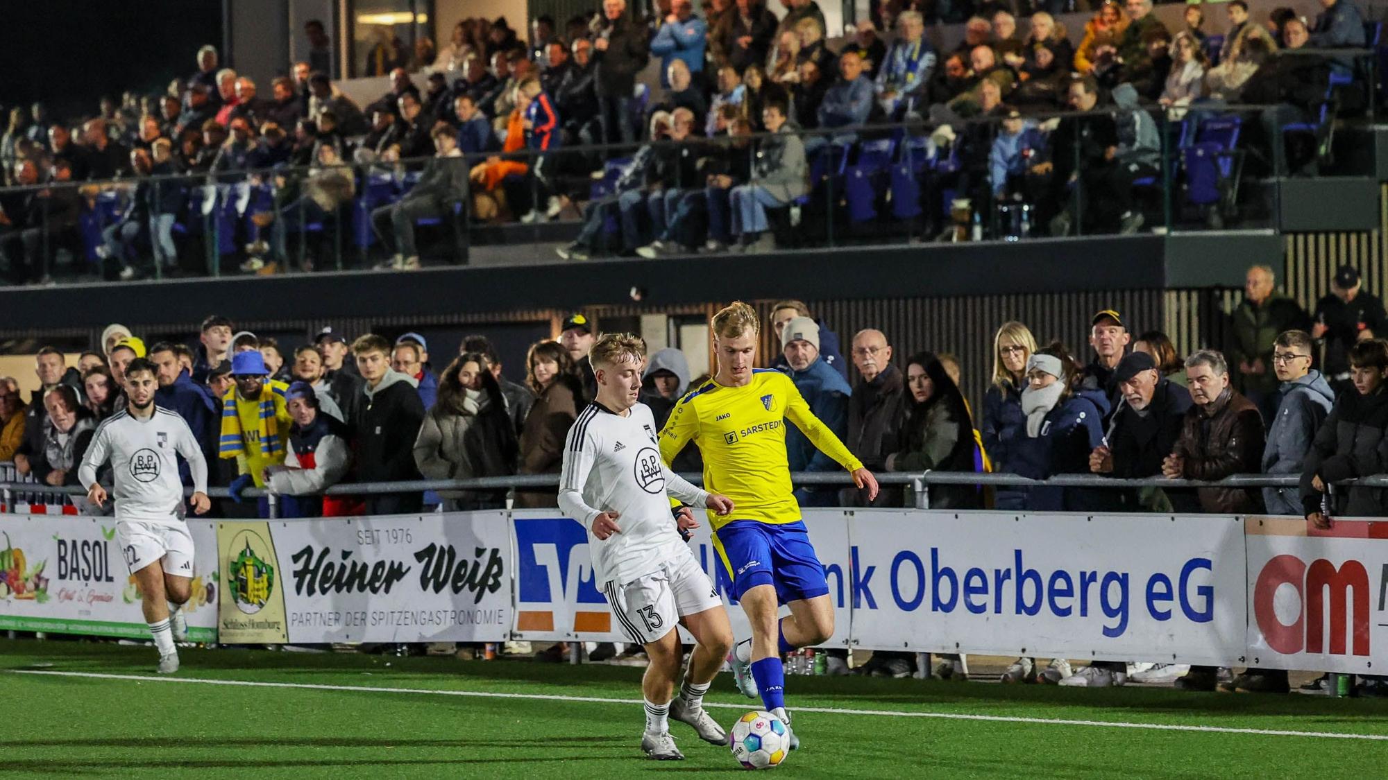 Vor voller Tribüne laufen ein Wiehler und ein Nümbrechter Fußballer um den Ball.