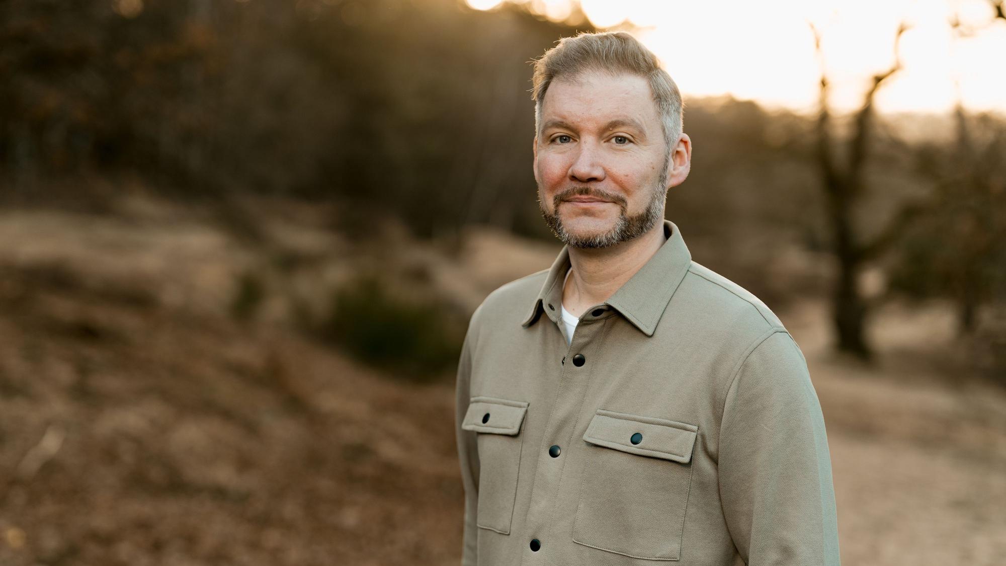 Der Autor Torsten Woywod steht in der Natur, die Landschaft ist nur unscharf und zu erahnen.