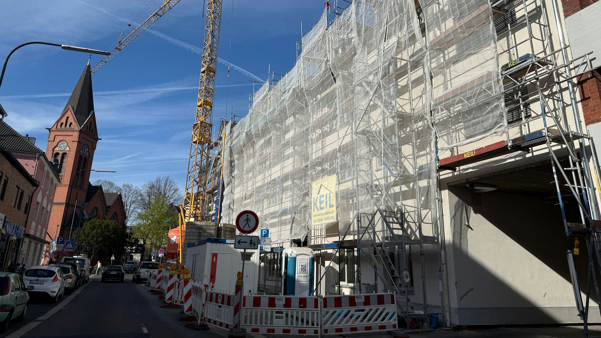 Eine Baustelle, Häuser hinter einem Schutznetz, ein Baukran und ein Kirchturm.
