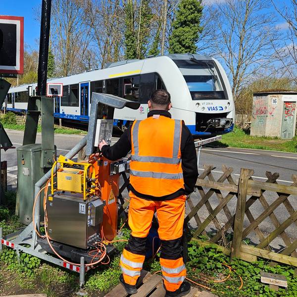 Ein Mitarbeiter der Bahn regelt den Einsatz einer Schranke an einem Bahnübergang, ein Zug fährt vorbei.