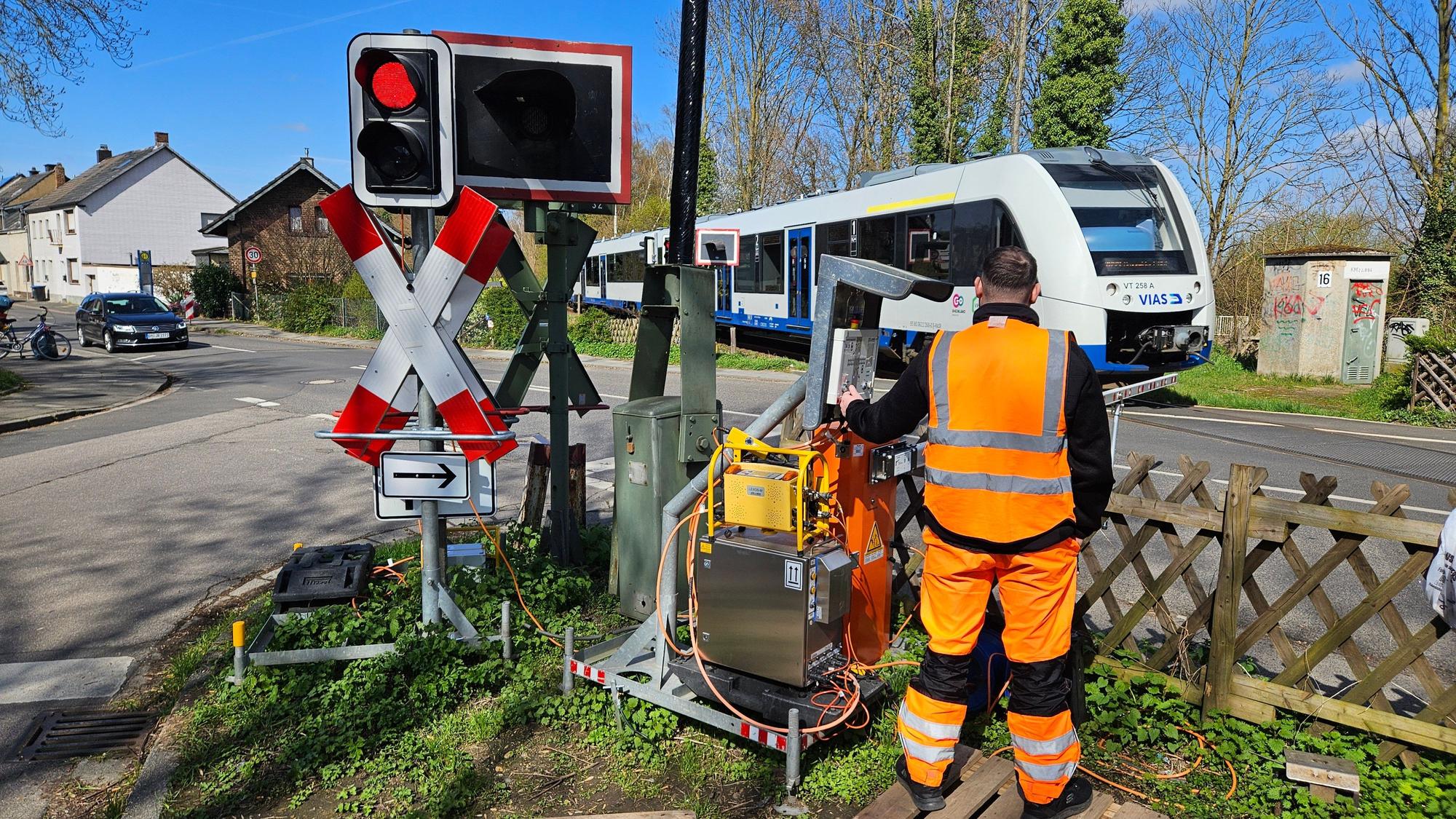 Ein Mitarbeiter der Bahn regelt den Einsatz einer Schranke an einem Bahnübergang, ein Zug fährt vorbei.