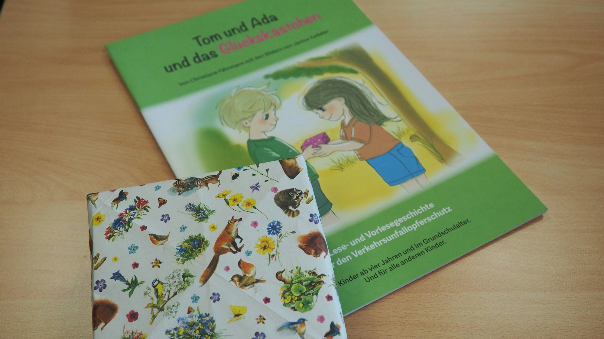 Das Buch „Tom und Ada und das Glückskästchen“ liegt auf einem Tisch, ebenso ein Kästchen aus buntem Geschenkpapier.