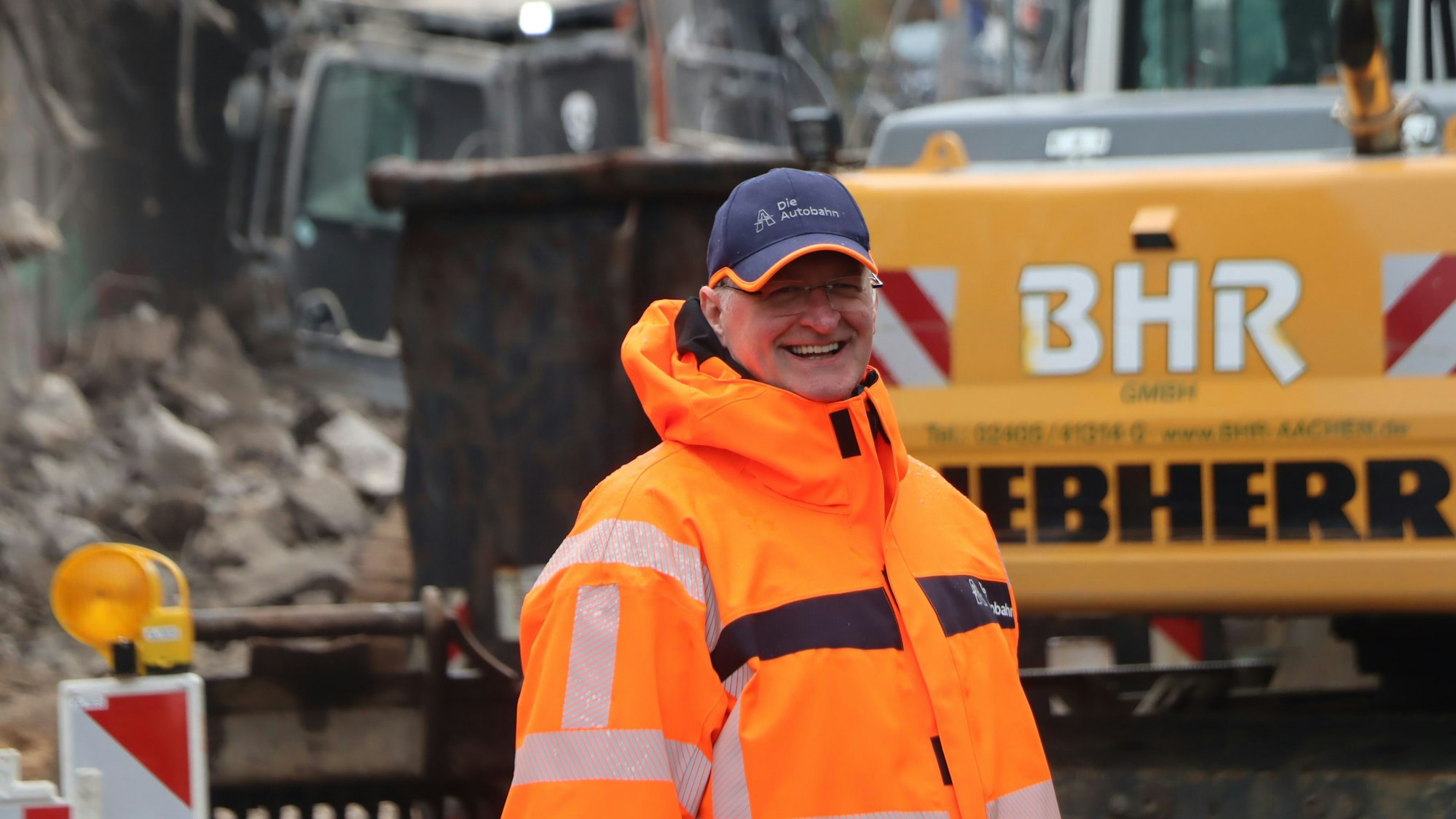 Auf dem Bild ist ein Mann in einer organgefarbenen Warnjacke auf einer Baustelle zu sehen.