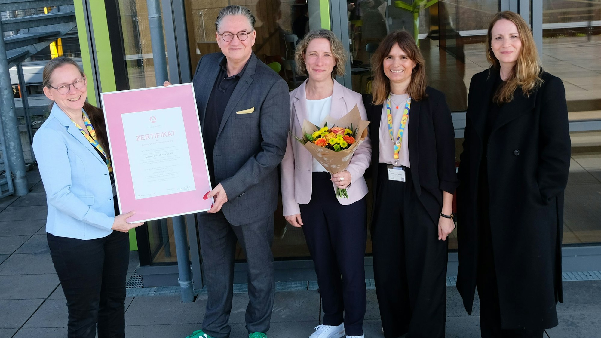 Fünf Personen stehen vor einem Gebäude, halten ein Zertifikat hoch und lächeln.