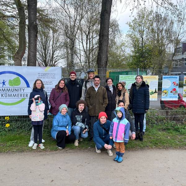 Eine Gruppe von Menschen steht vor einem Zaun. Im Hintergrund hängt ein Banner am Zaun mit der Überschrift Community der Kümmerer.