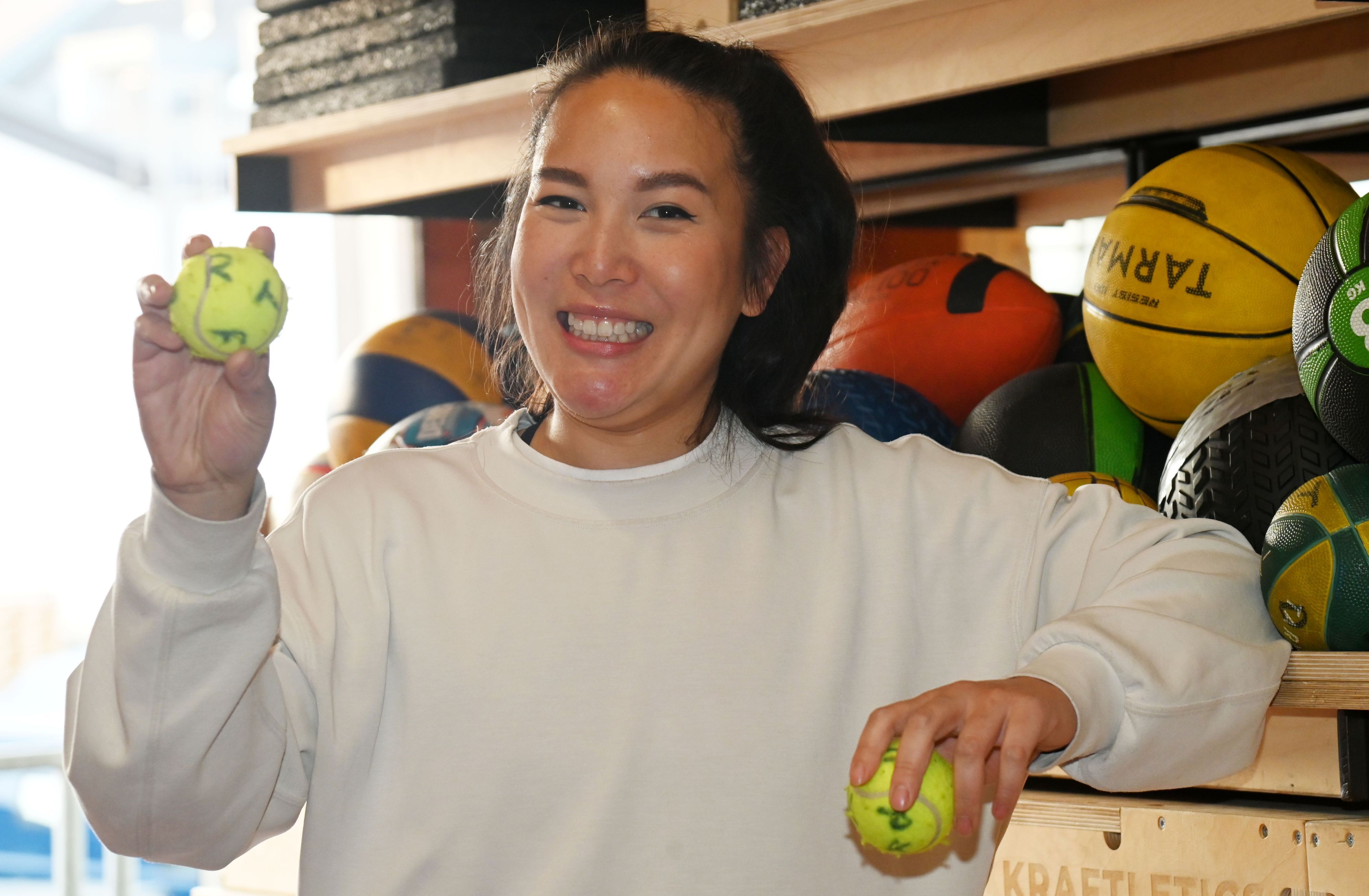 Tina Nguyen steht vor einem Regal mit Bällen. Sie lächelt und hält einen beschrifteten Tennisball in der Hand