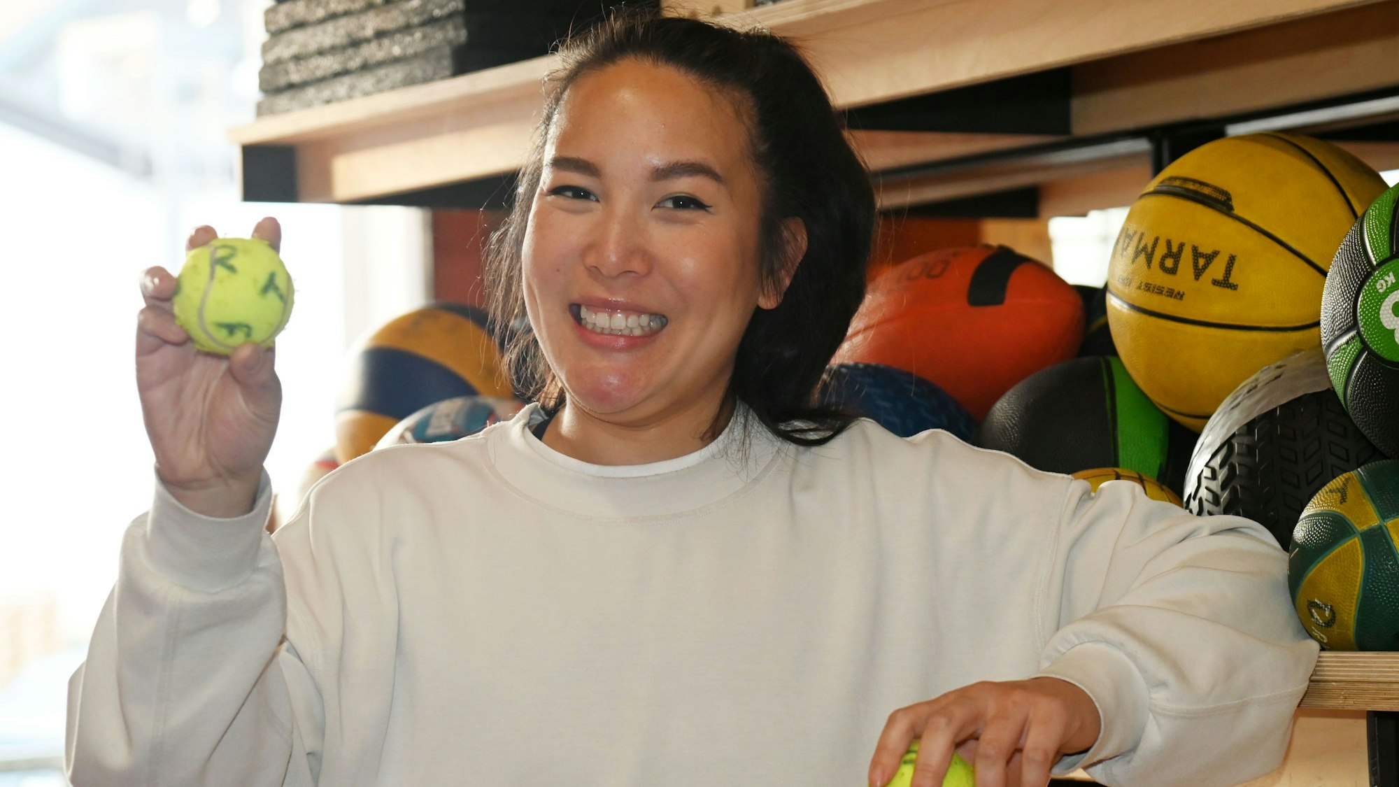 Tina Nguyen steht vor einem Regal mit Bällen. Sie lächelt und hält einen beschrifteten Tennisball in der Hand