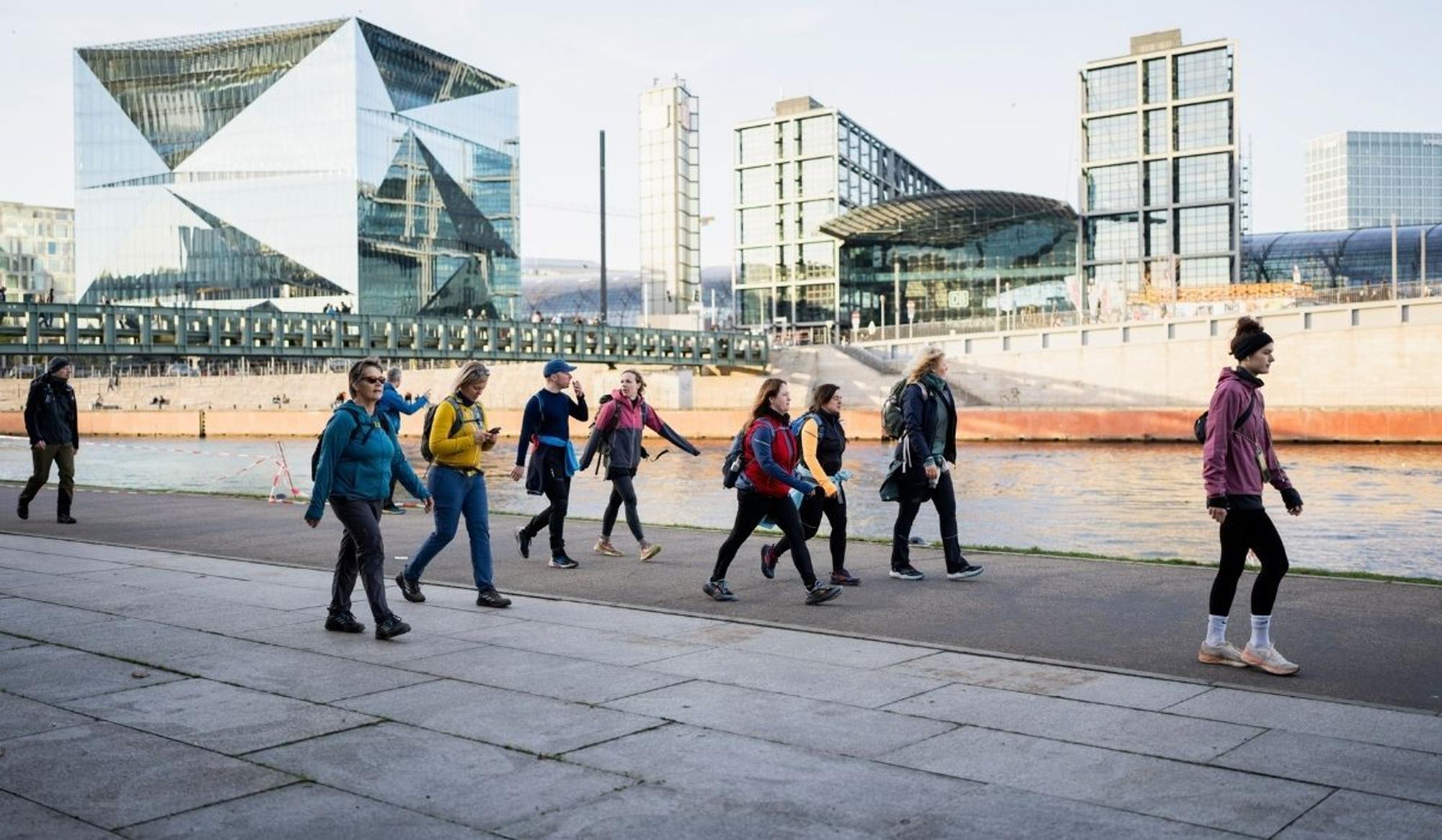 Gruppe von Mammutmarsch-Teilnehmenden in bunter Outdoor-Kleidung und mit Rucksäcken, die entlang eines Uferwegs vor einer modernen Stadtsilhouette mit Glasfassaden und einem Bahnhof wandern – aufgenommen bei kühlem, klarem Wetter.