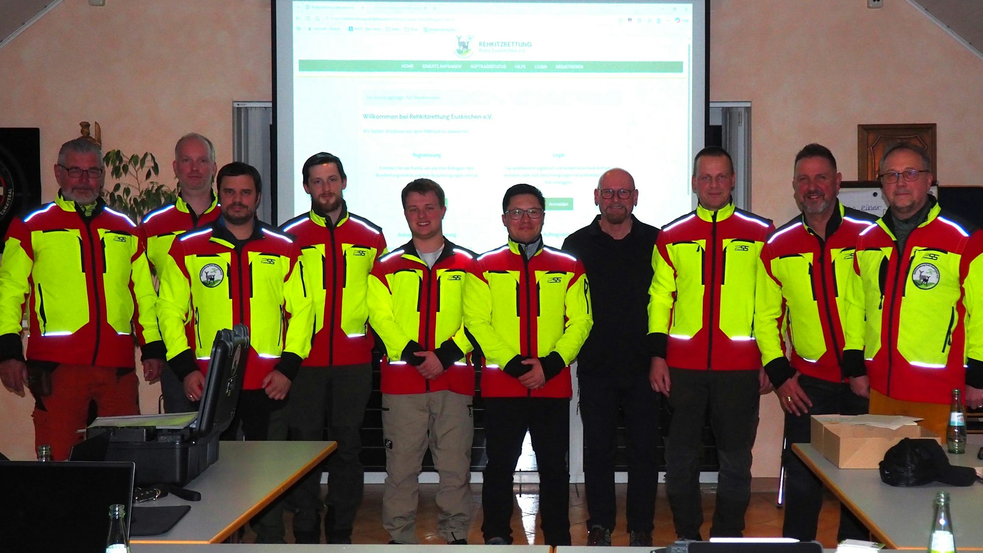 Die zehn Drohnenpiloten stehen vor einer Leindwand, auf der das neue Buchungssystem gezeigt wird. Die meisten haben neongelbe Jacken an.