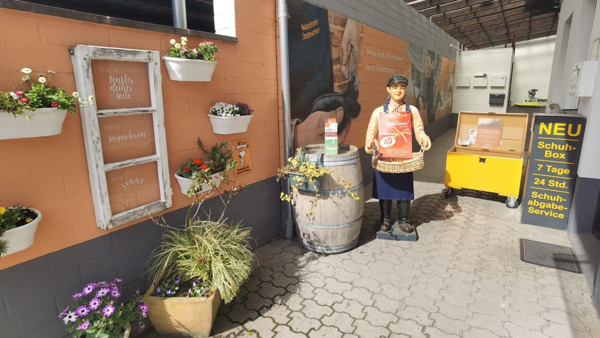 Zu sehen ist der Außenbereich einer Schuhmacherei mit einer Werbefigur, Blumen und einer Box für Schuhe, die gespendet werden.