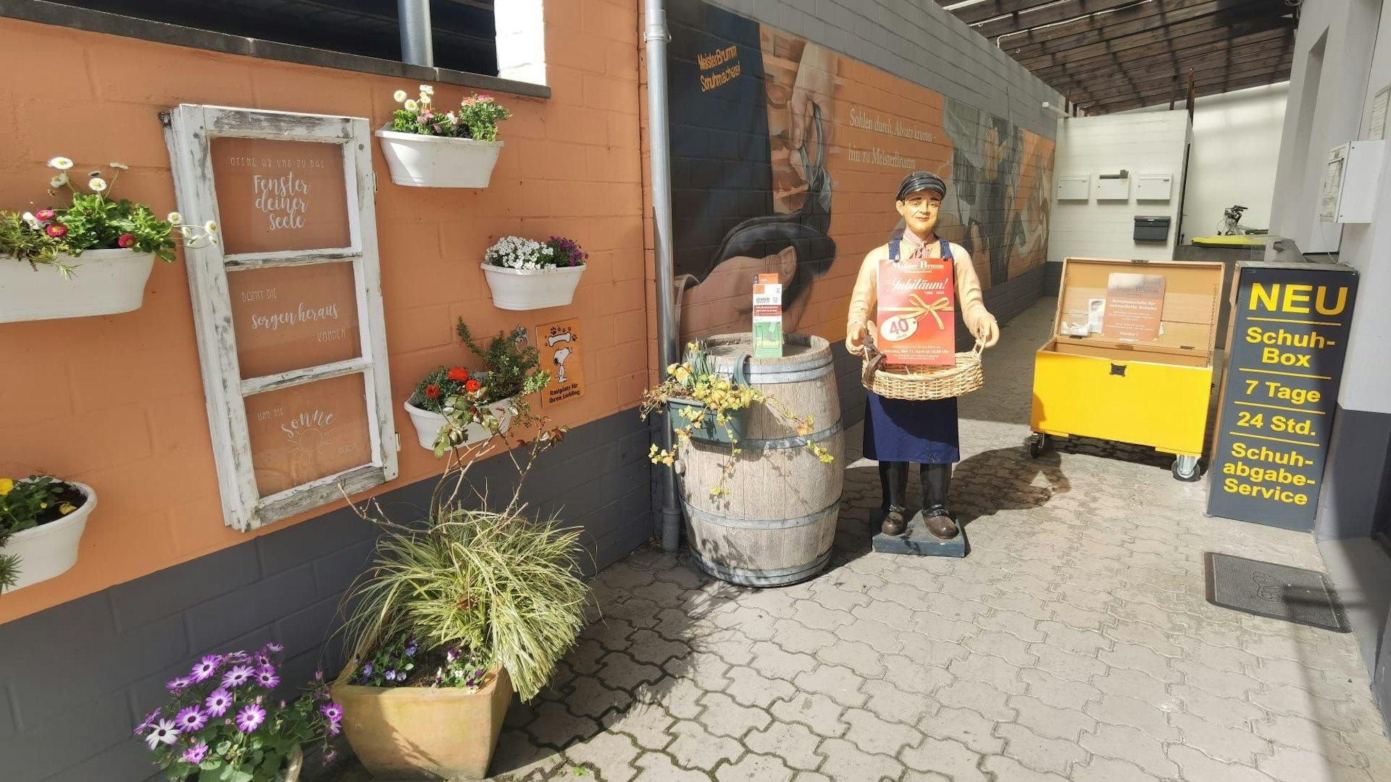 Zu sehen ist der Außenbereich einer Schuhmacherei mit einer Werbefigur, Blumen und einer Box für Schuhe, die gespendet werden.