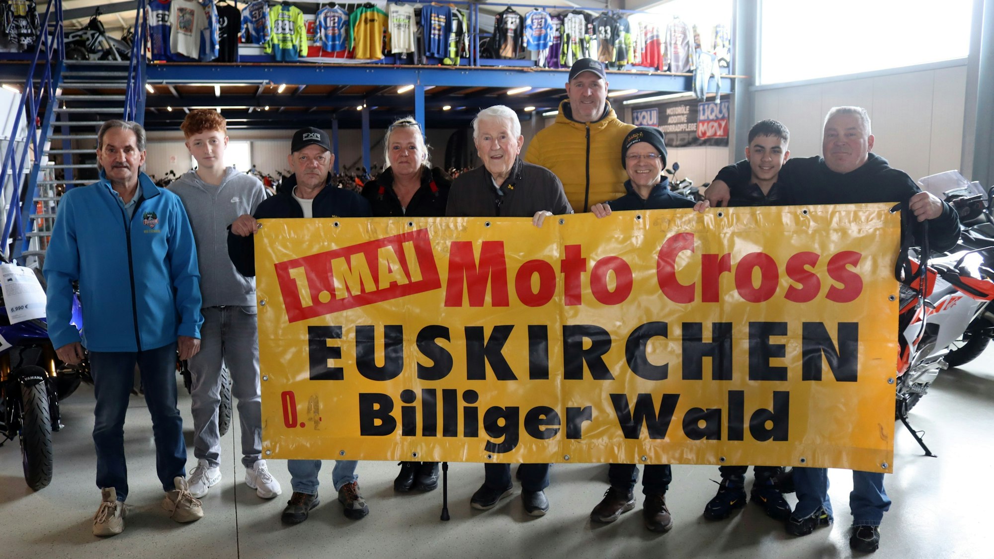 Acht Männer und eine Frau stehen in einem Motorradfachhandel und halten ein Plakat hoch, das die Motocrossveranstaltugn am 1. Mai im Billiger Wald bewirbt.