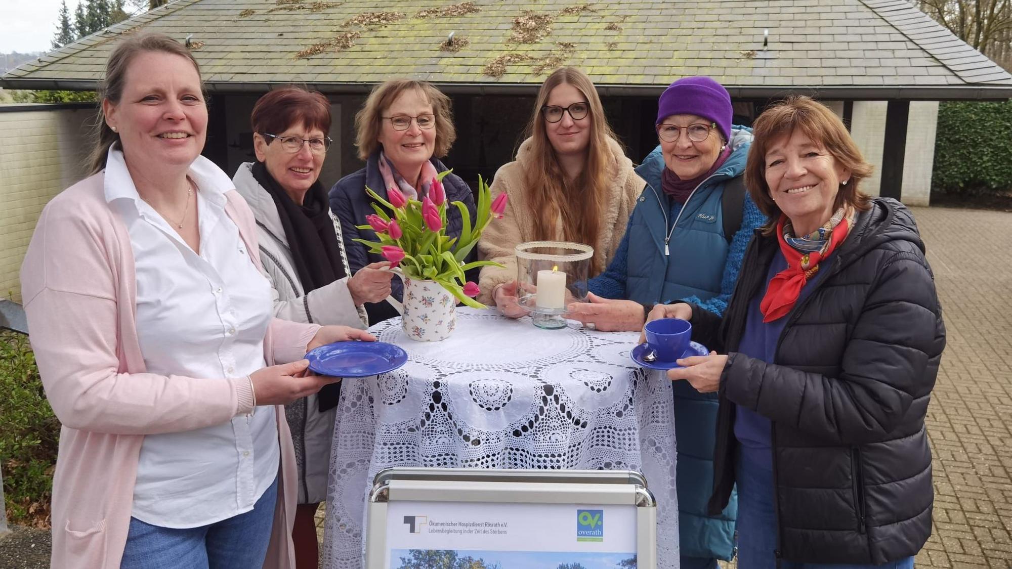 Eine Gruppe Frauen steht um einen Stehtisch herum. Sie haben Tassen und Kuchenteller in der Hand und lächeln.
