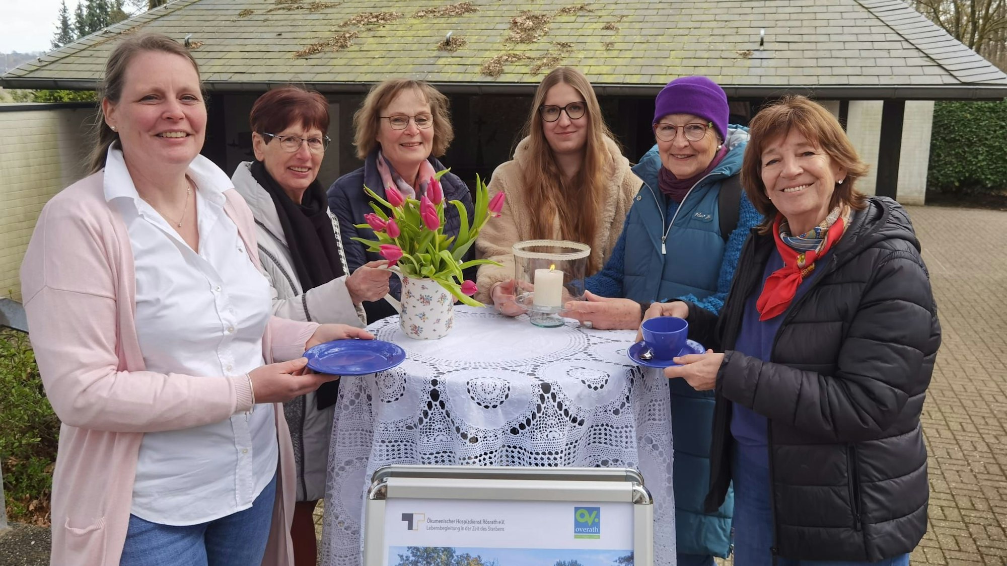 Eine Gruppe Frauen steht um einen Stehtisch herum. Sie haben Tassen und Kuchenteller in der Hand und lächeln.