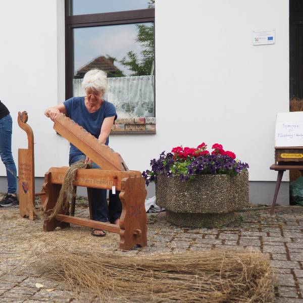 Zwei Frauen bearbeiten Flachshalme mit historischen Werkzeugen, rechts im Bild steht ein Blumenkübel.