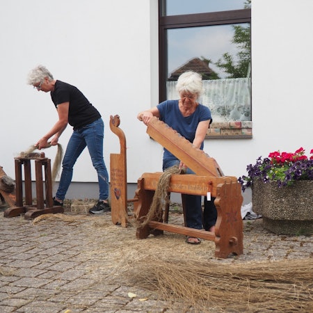 Zwei Frauen bearbeiten Flachshalme mit historischen Werkzeugen, rechts im Bild steht ein Blumenkübel.