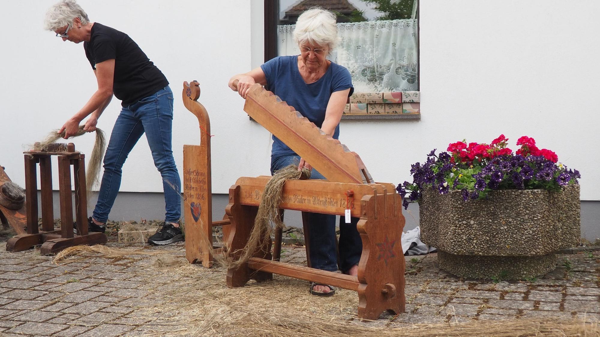 Zwei Frauen bearbeiten Flachshalme mit historischen Werkzeugen, rechts im Bild steht ein Blumenkübel.