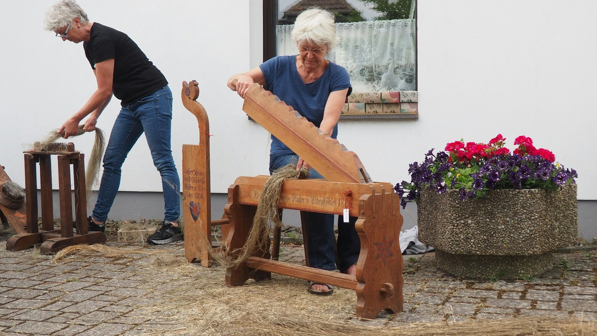 Zwei Frauen bearbeiten Flachshalme mit historischen Werkzeugen, rechts im Bild steht ein Blumenkübel.