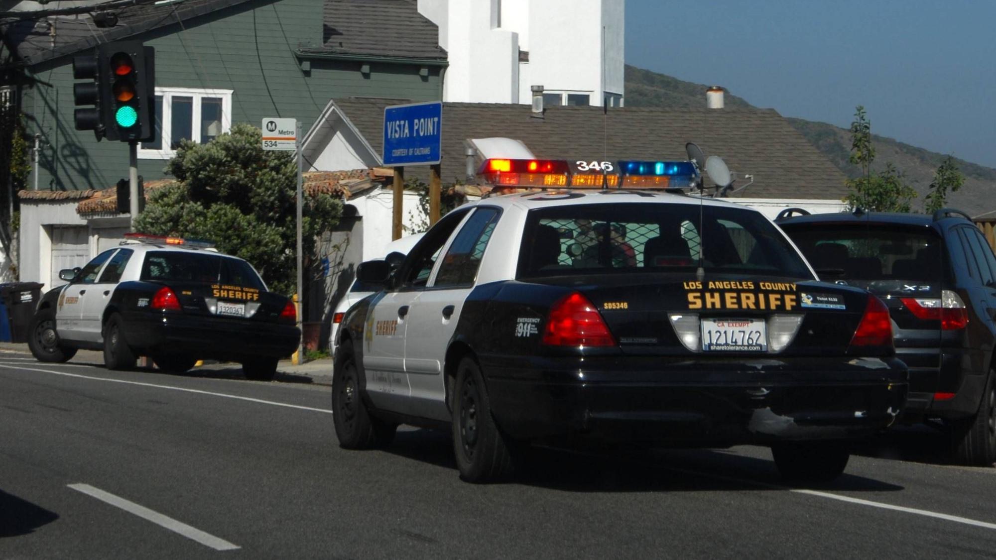Zwei „Los Angeles County Sheriff“ Autos auf einer Straße mit Häusern im Hintergrund.