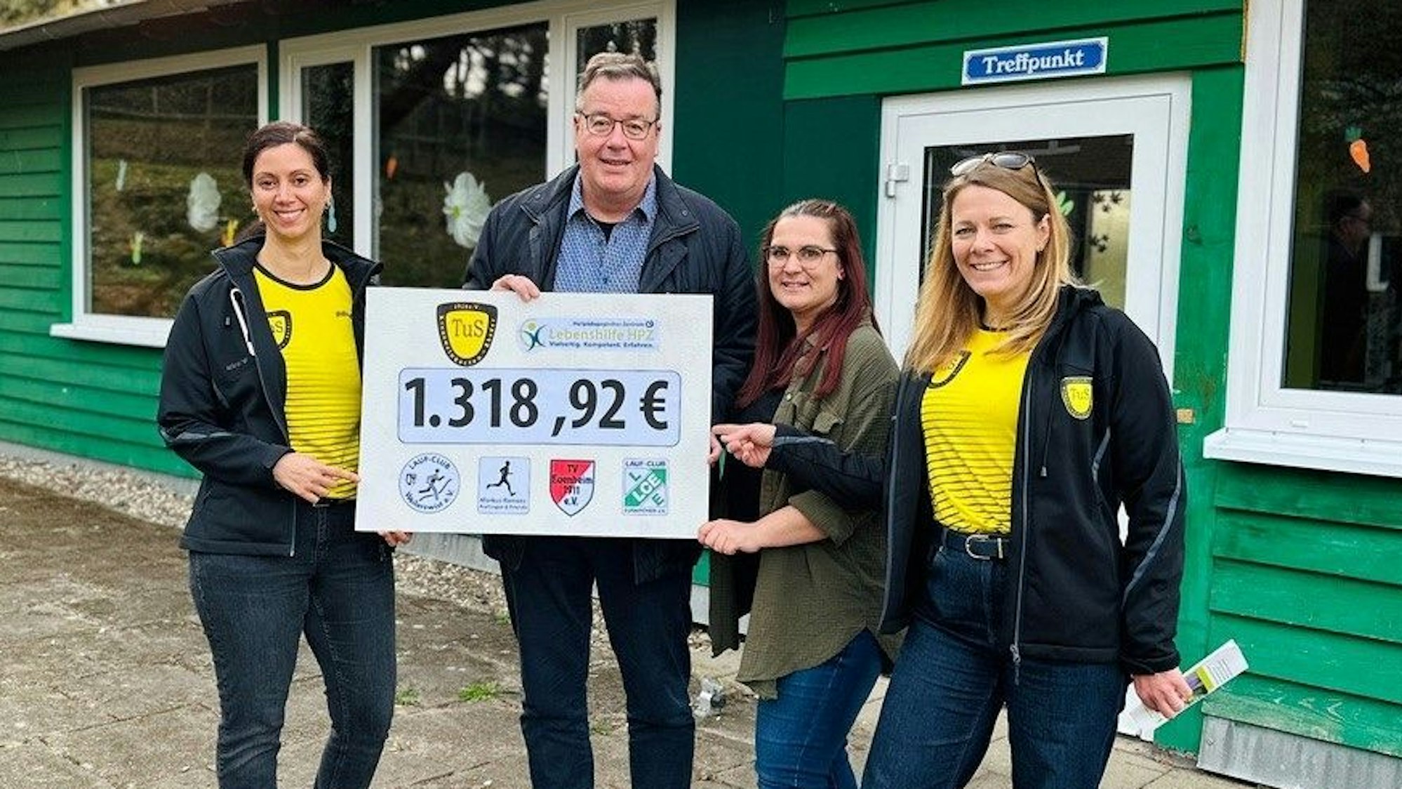 Zwei Vertreterinnen des TuS Kreuzweingarten-Rheder in gelben Trikots stehen mit einem Mann und einer Frau vor einer grünen Blockhütte.