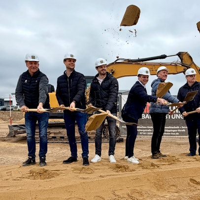 Sechs Männer stehen vor einem Bagger und werfen mit Schaufeln Sand in die Luft.