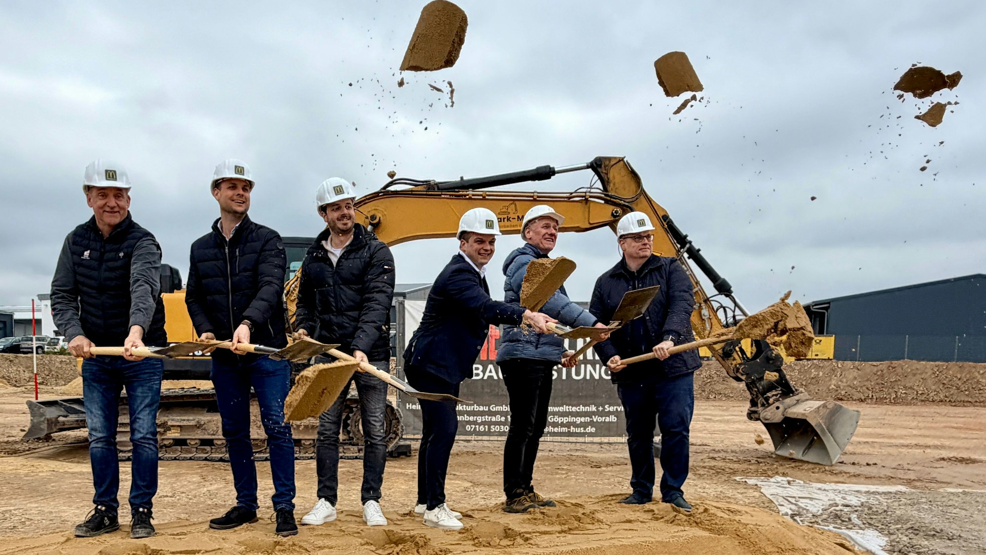 Sechs Männer stehen vor einem Bagger und werfen mit Schaufeln Sand in die Luft.