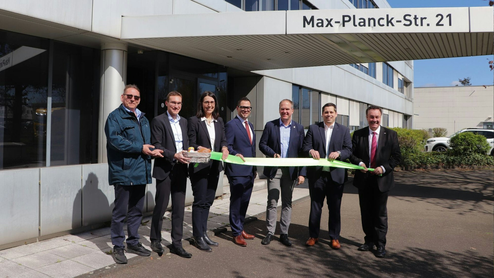 Das Foto zeigt eine Gruppe von Menschen mit grünem Band und einem Präsentkorb vor dem Gebäude der neuen Niederlassung.