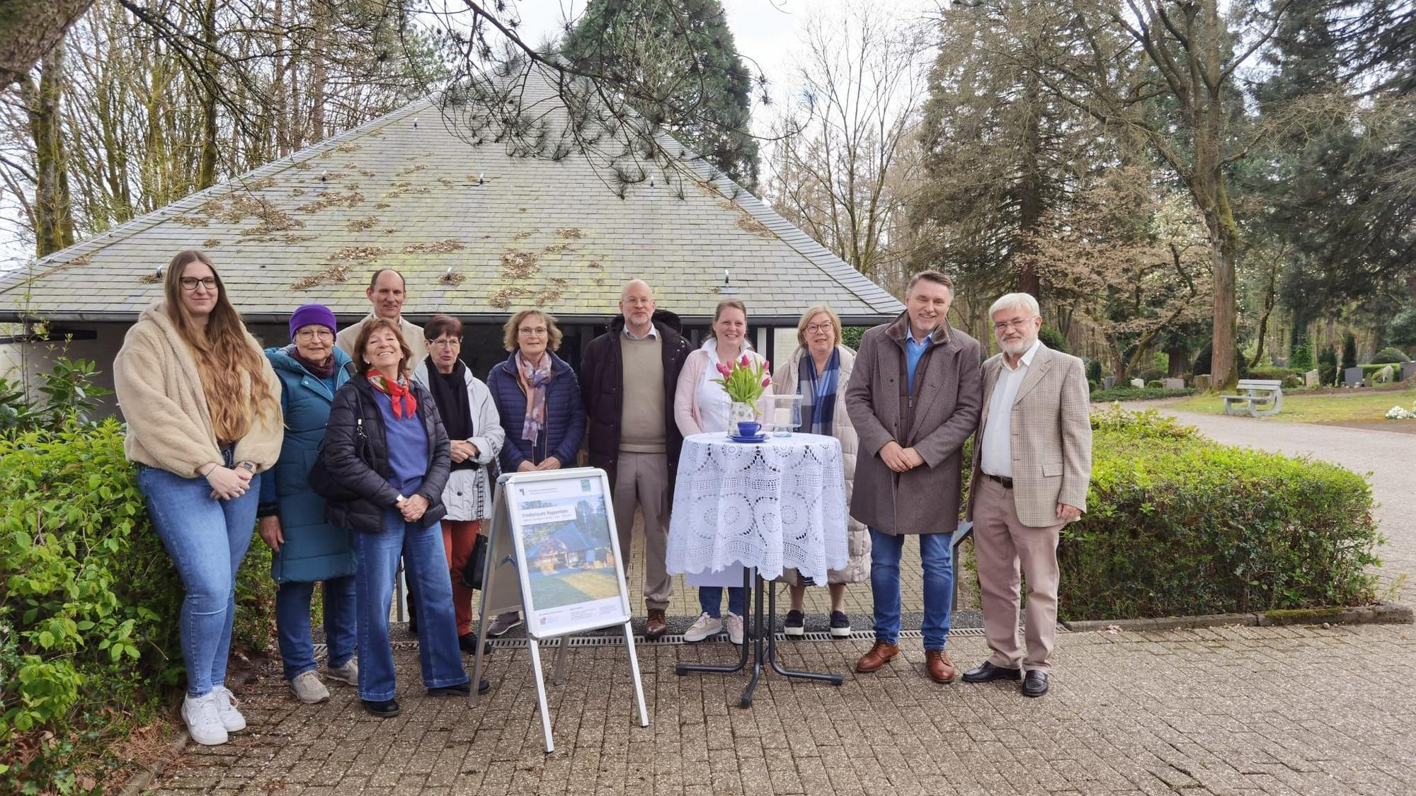 Eine Gruppe steht auf einem Friedhof vor einer Trauerhalle und lächelt.