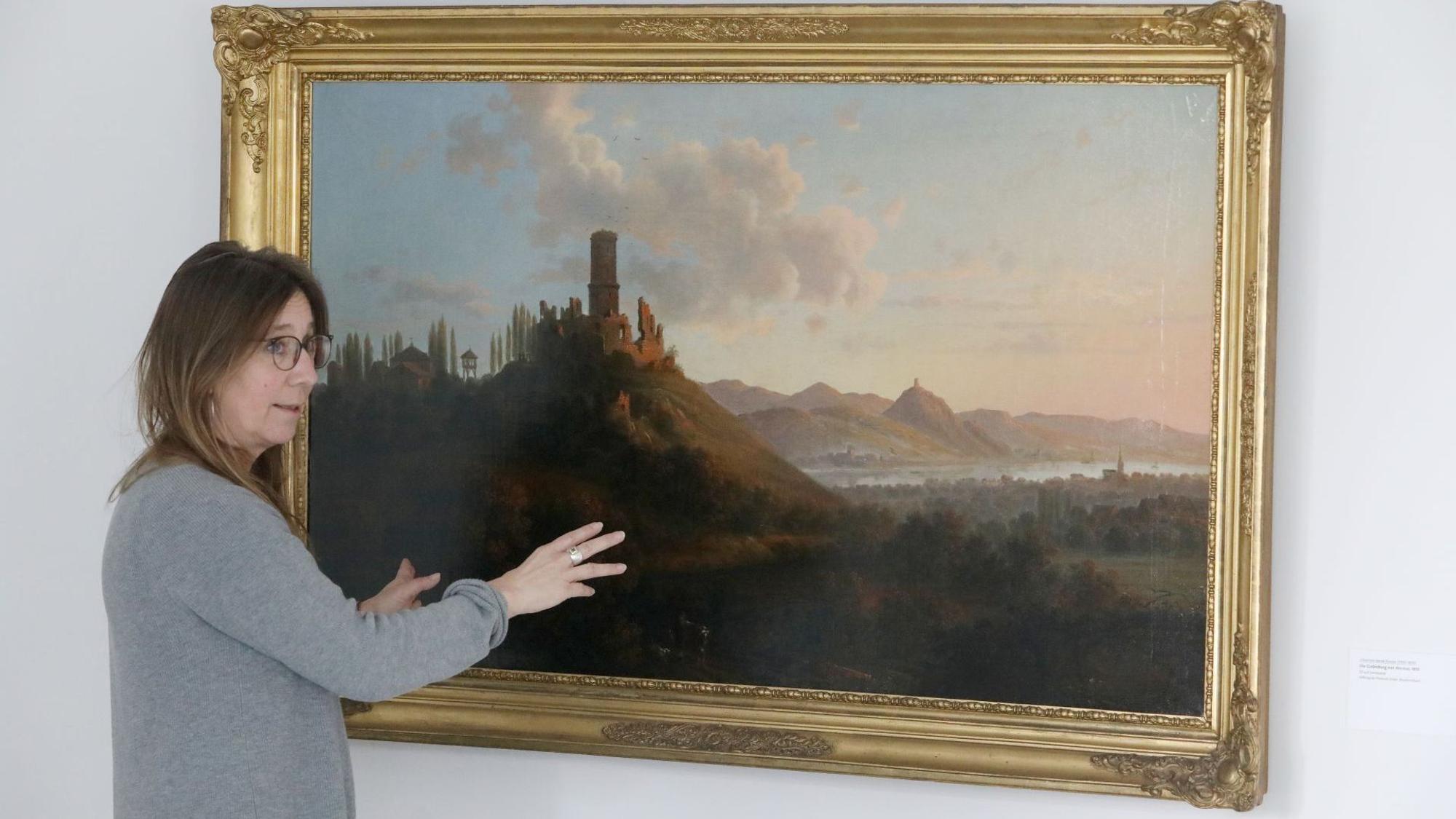Eine Frau vor einem historischen Ölgemälde mit der Darstellung einer Rheinlandschaft.