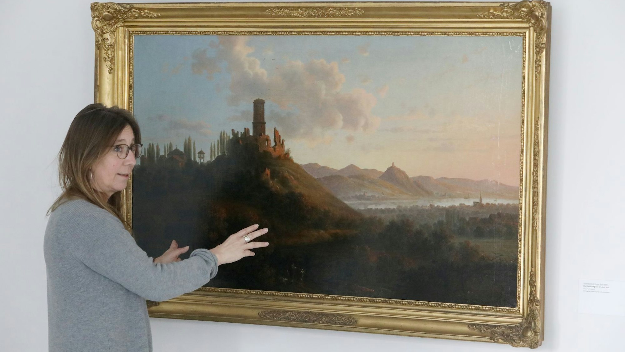 Eine Frau vor einem historischen Ölgemälde mit der Darstellung einer Rheinlandschaft.