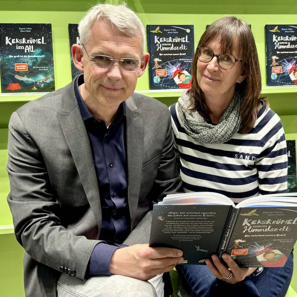 Eine Frau und ein Mann lächeln in die Kamera. Im Hintergrund sind Bücher in einem Regal zu sehen.