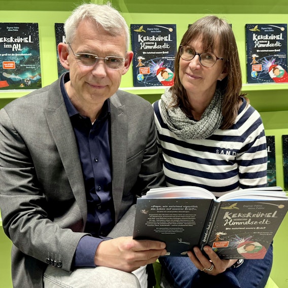 Eine Frau und ein Mann lächeln in die Kamera. Im Hintergrund sind Bücher in einem Regal zu sehen.