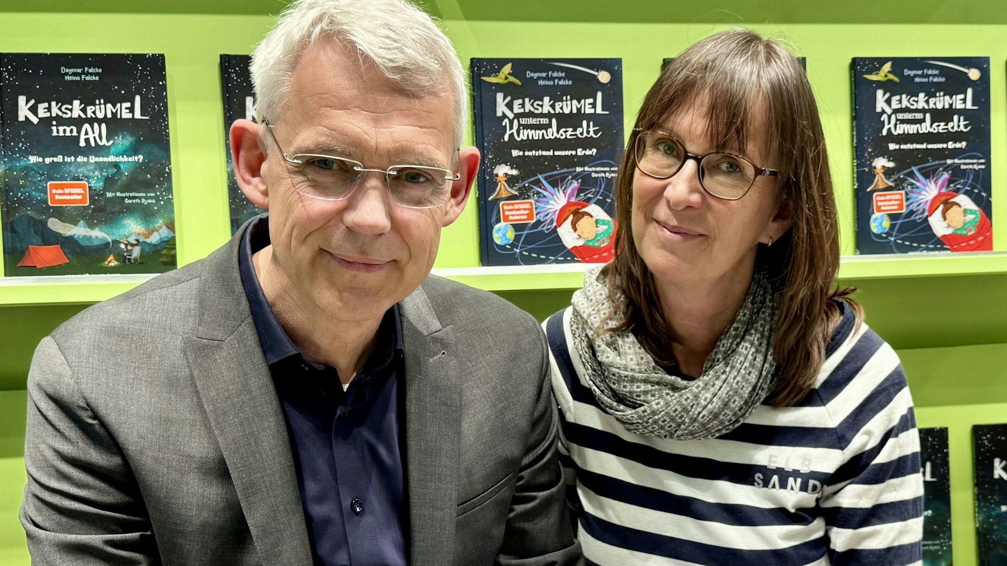 Eine Frau und ein Mann lächeln in die Kamera. Im Hintergrund sind Bücher in einem Regal zu sehen.