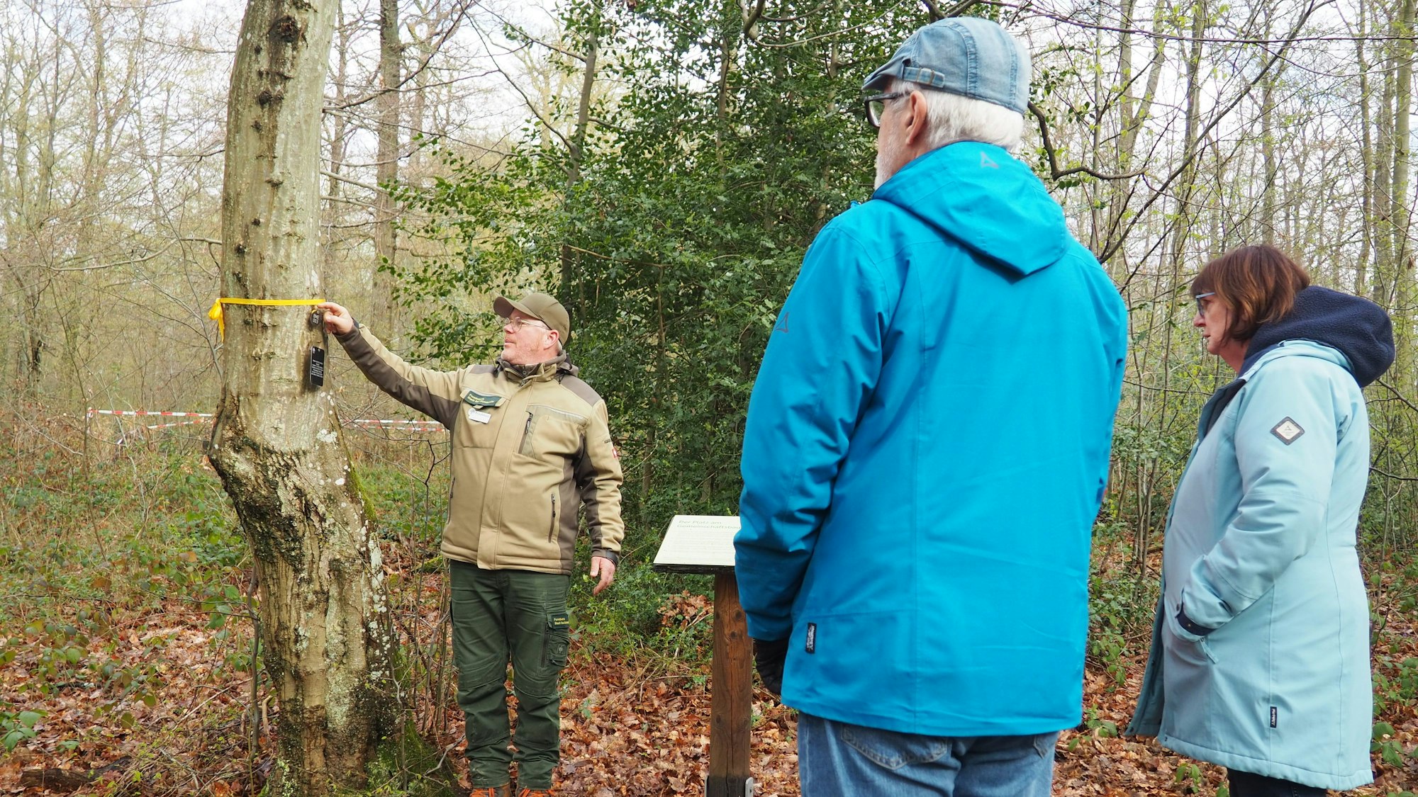 Frank Hallmanns zeigt auf ein gelbes Band an einem Baum. Ein Mann und eine Frau, von hinten zu sehen, schauen in seine Richtung.