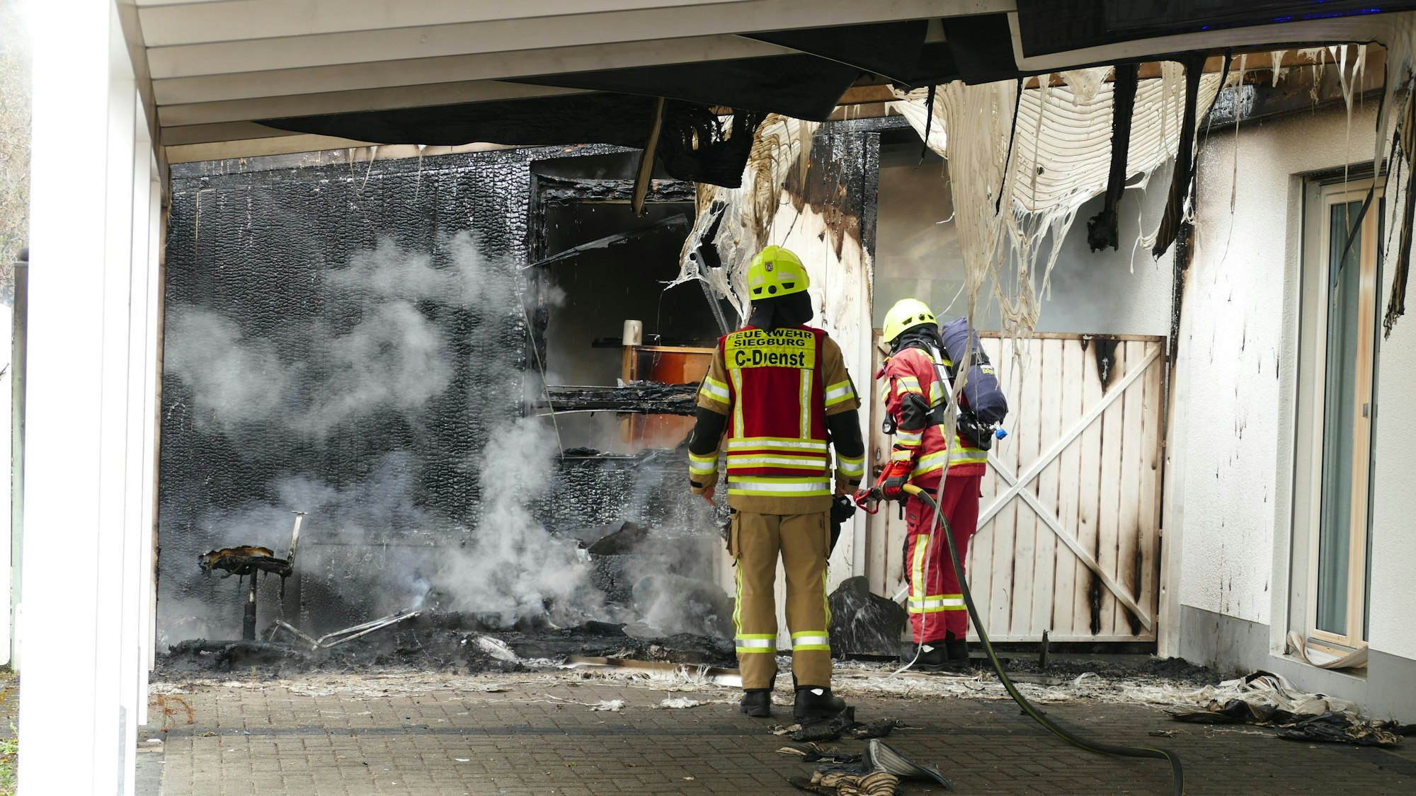 Feuerwehrleute löschen das Feuer in einem Schuppen an einer Doppelhaushälfte. Die Wände des Schuppens sind komplett verkohlt.