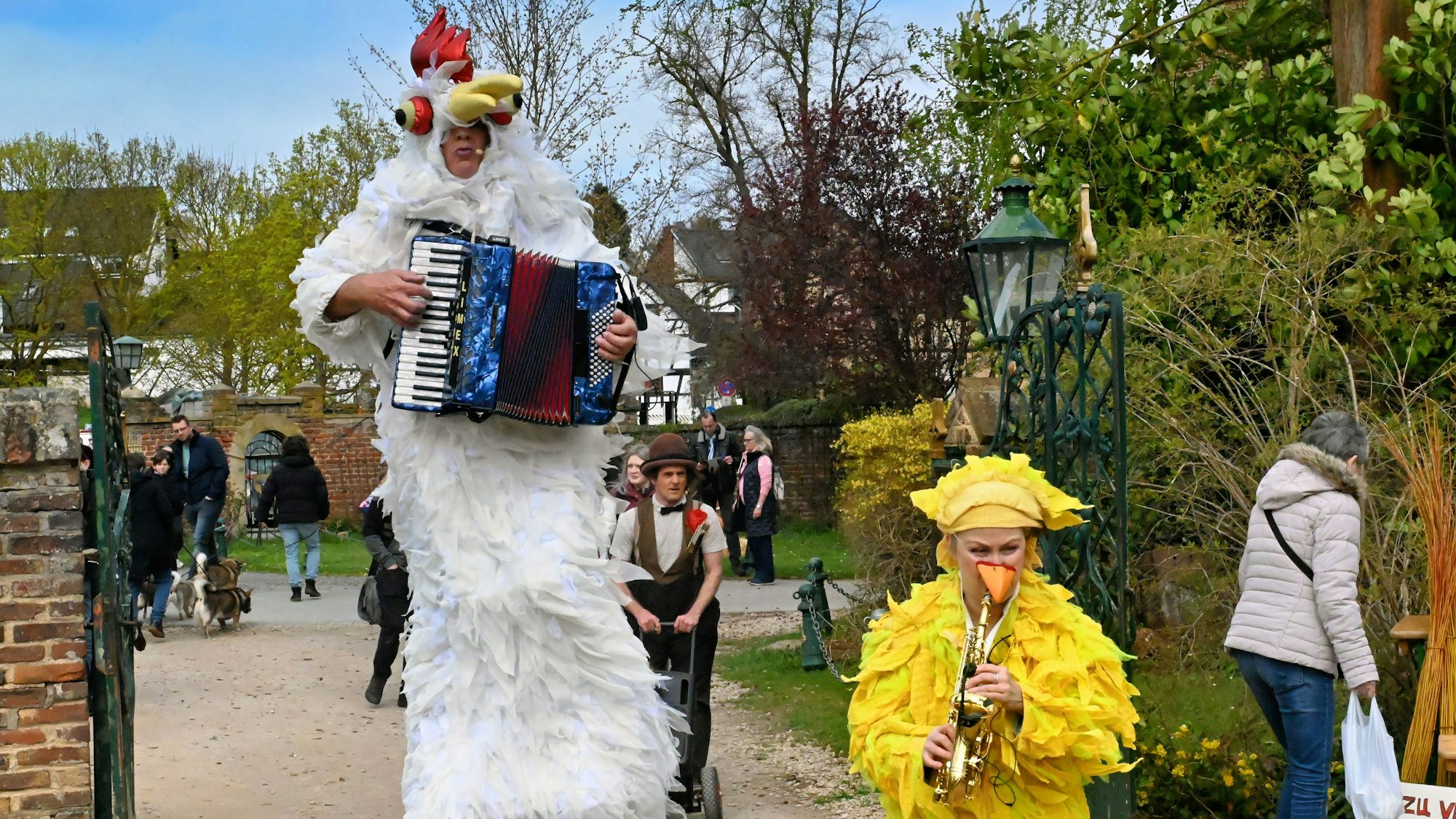 Auf Stelzen läuft ein als wißes Huhn verkleideter Akkordeonspieler übers Gelände, vor ihm, nur halb so groß, eine Musikerkollegin im gelben Federkleid.