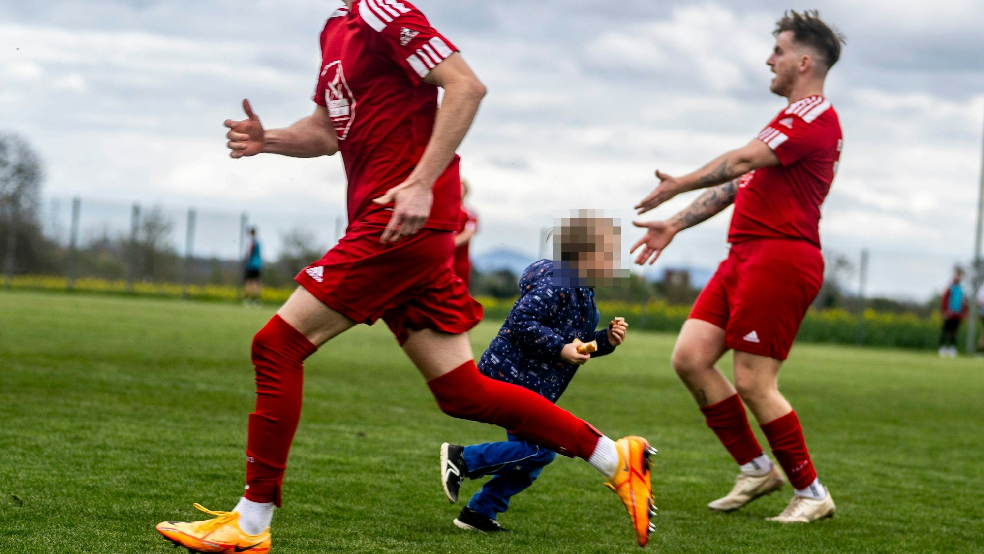 Ein Kind läuft, mit der Wurst in der Hand, übers Spielfeld und zwischen zwei Verteidigern hindurch. Einer von ihnen weist gestenreich auf den Flitzer hin.