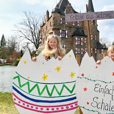 Die beiden Kinder stehen hinter zwei großen Pappeiern. Auf einem steht zu lesen: „Einfach mal in Schale werfen“. Hinter ihnen ein Schild mit der Aufschrift „Grüße aus Satzvey“, dahinter die Burg.